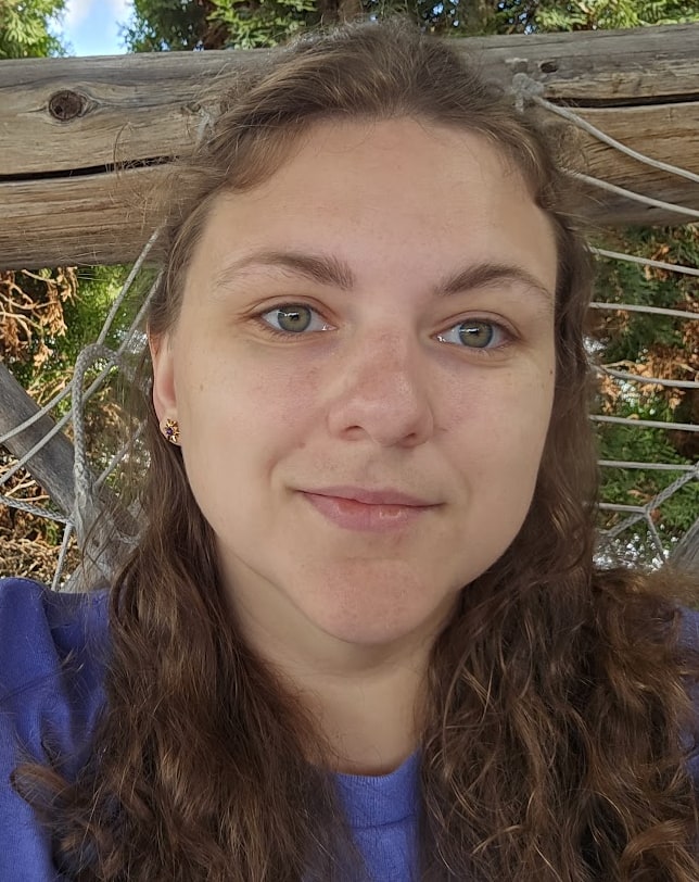 Katherine Dattilo, smiling, outdoors in front of a wooden trellis and evergreens.